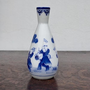 May include: White porcelain vase with a blue and white design. The vase has a narrow neck and a bulbous body. The blue design depicts figures in a playful scene. The rim of the vase has a blue decorative pattern.
