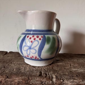 May include: A white ceramic pitcher with a blue, green, and red floral design. The pitcher has a handle and a spout. The base is white, and the top is a lighter shade of white. The pitcher is sitting on a wooden surface.