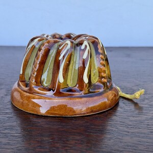 May include: A brown ceramic pudding mold with a decorative design. The mold features vertical stripes in shades of brown, green, and white. A yellow string is attached to the side. The mold is likely vintage and suitable for baking.