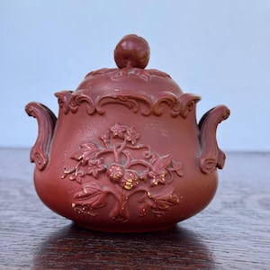 Può includere: Una teiera in ceramica color marrone con coperchio e due manici. La teiera è decorata con un motivo floreale e un piccolo pomello sul coperchio. La teiera è appoggiata su una superficie di legno.