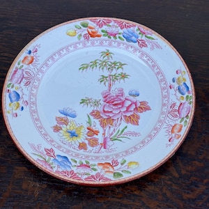 May include: A white ceramic plate with a floral design. The plate features a central image of a pink peony and a tree, surrounded by colorful flowers. The rim has a pink border with a repeating pattern. The plate is approximately 8 inches in diameter.