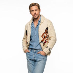 May include: A cream-coloured, chunky knit cardigan with a shawl collar. The sweater features a large dog face design in brown, black, and red on the front and paw prints on the cuffs. The person is wearing a blue denim shirt and light blue jeans.