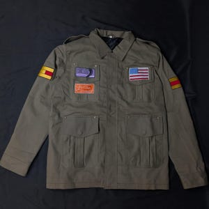May include: An olive green utility jacket featuring four front pockets, shoulder epaulets, and a central zip closure. The chest displays a rectangular patch with purple text, an orange patch with white text, and a United States flag patch.