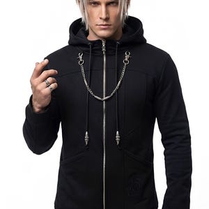 May include: A black zip-up hoodie featuring a silver-toned metal chain draped across the chest, attached to the collar with lobster clasps. The garment includes a hood with drawstrings, long sleeves, and a subtle embossed logo on the lower front.