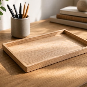 Könnte beinhalten: Ein rechteckiges Holztablett mit erhöhten Kanten liegt auf einer Holzoberfläche. Ein kleiner Blumentopf, ein Keramik-Stifthalter mit Stiften und Bleistiften sowie ein Stapel Bücher befinden sich ebenfalls auf dem Tisch. Das Tablett ist aus hellem Holz.