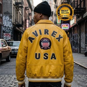 May include: A mustard yellow leather varsity jacket featuring the text AVIREX in black arched lettering across the upper back, with the word USA below and a central circular patch displaying an American flag and the text ALL AMERICAN 1975.