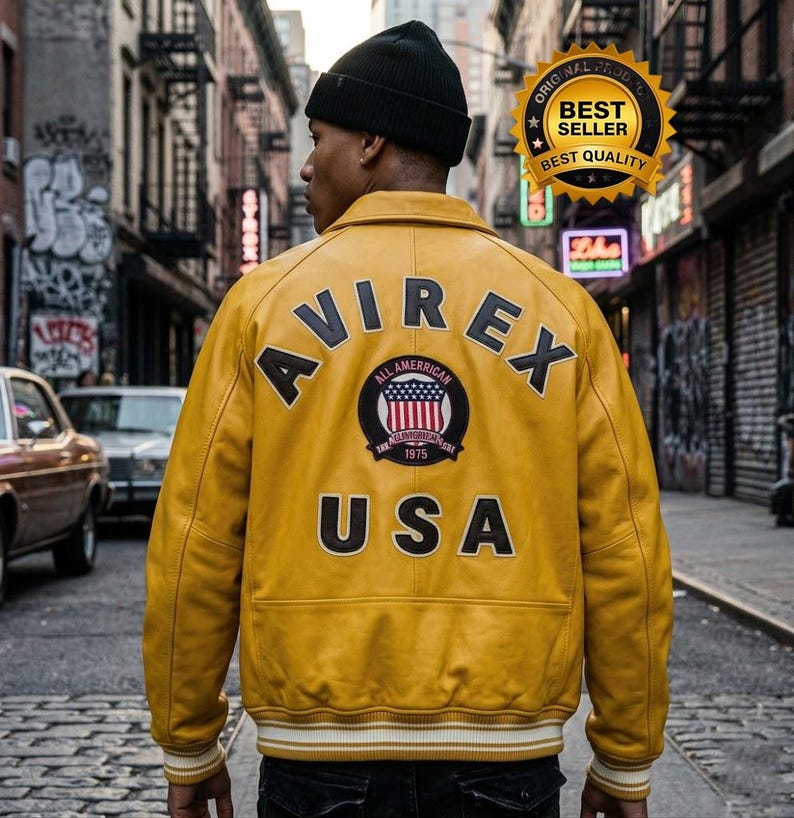 May include: A mustard yellow leather varsity jacket featuring the text AVIREX in black arched lettering across the upper back, with the word USA below and a central circular patch displaying an American flag and the text ALL AMERICAN 1975.