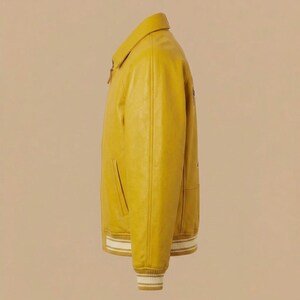 May include: A mustard yellow jacket with a classic collar, a front zipper closure, and long sleeves. The cuffs and the bottom hem feature a ribbed knit pattern with white stripes. The jacket has two side pockets and a smooth, solid-colored finish.