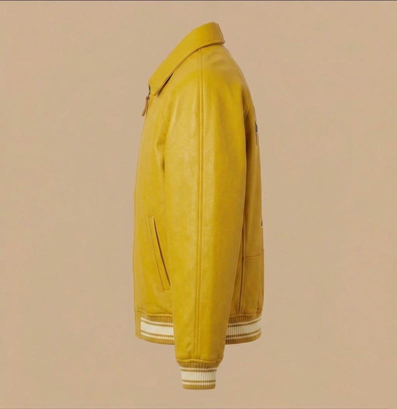 May include: A mustard yellow jacket with a classic collar, a front zipper closure, and long sleeves. The cuffs and the bottom hem feature a ribbed knit pattern with white stripes. The jacket has two side pockets and a smooth, solid-colored finish.