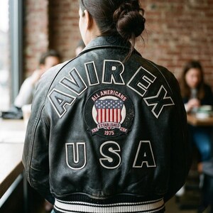 Puede incluir: Chaqueta de cuero negra con letras blancas que deletrean "AVIREX" y "USA". Un parche circular presenta la bandera estadounidense y las palabras "All Americans" y "Established 1975". La chaqueta tiene un dobladillo a rayas.