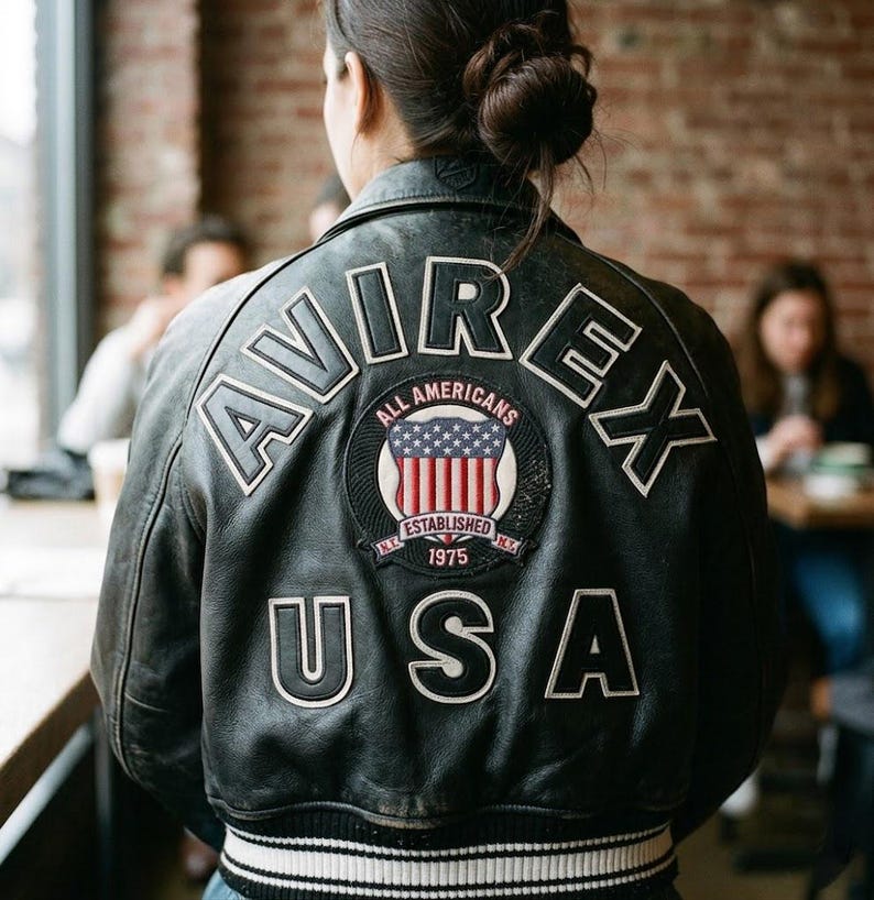 Puede incluir: Chaqueta de cuero negra con letras blancas que deletrean "AVIREX" y "USA". Un parche circular presenta la bandera estadounidense y las palabras "All Americans" y "Established 1975". La chaqueta tiene un dobladillo a rayas.