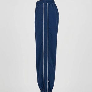 May include: A pair of navy blue track pants featuring a white vertical stripe running down the side of each leg. The pants have an elastic waistband and elastic cuffs at the ankles, with a small rectangular logo tag near the bottom hem.