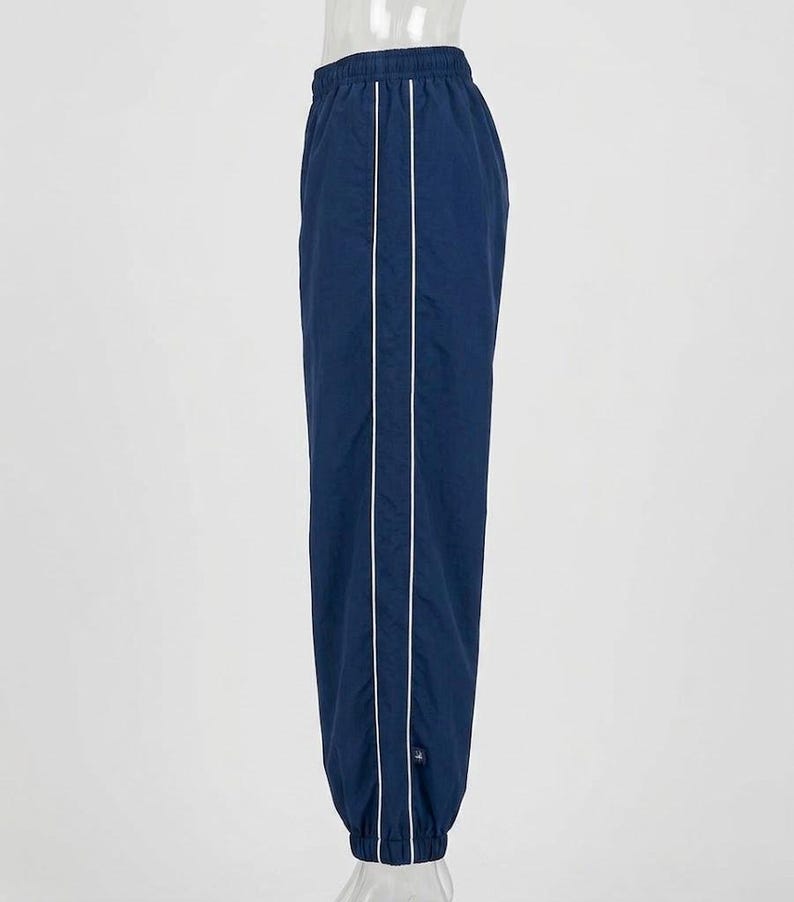 May include: A pair of navy blue track pants featuring a white vertical stripe running down the side of each leg. The pants have an elastic waistband and elastic cuffs at the ankles, with a small rectangular logo tag near the bottom hem.
