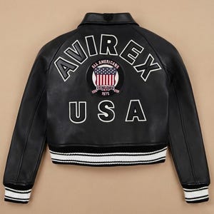 Puede incluir: Chaqueta de cuero negra con letras y parches bordados en blanco. La parte trasera de la chaqueta presenta la palabra "AVIREX" sobre un parche circular con un dise&ntilde;o de bandera estadounidense y la palabra "USA" debajo. Cuello y pu&ntilde;os acanalados.