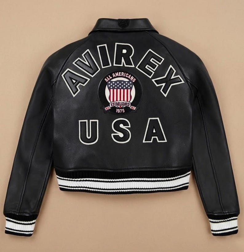 Puede incluir: Chaqueta de cuero negra con letras y parches bordados en blanco. La parte trasera de la chaqueta presenta la palabra "AVIREX" sobre un parche circular con un dise&ntilde;o de bandera estadounidense y la palabra "USA" debajo. Cuello y pu&ntilde;os acanalados.