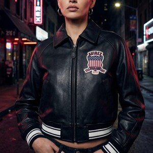 Puede incluir: Chaqueta bomber de cuero negro con cremallera, cuello y ribete de rayas blancas en los pu&ntilde;os y el dobladillo. Cuenta con un parche en el pecho con el texto "AVIREX" y un dise&ntilde;o de bandera estadounidense. La chaqueta es corta.