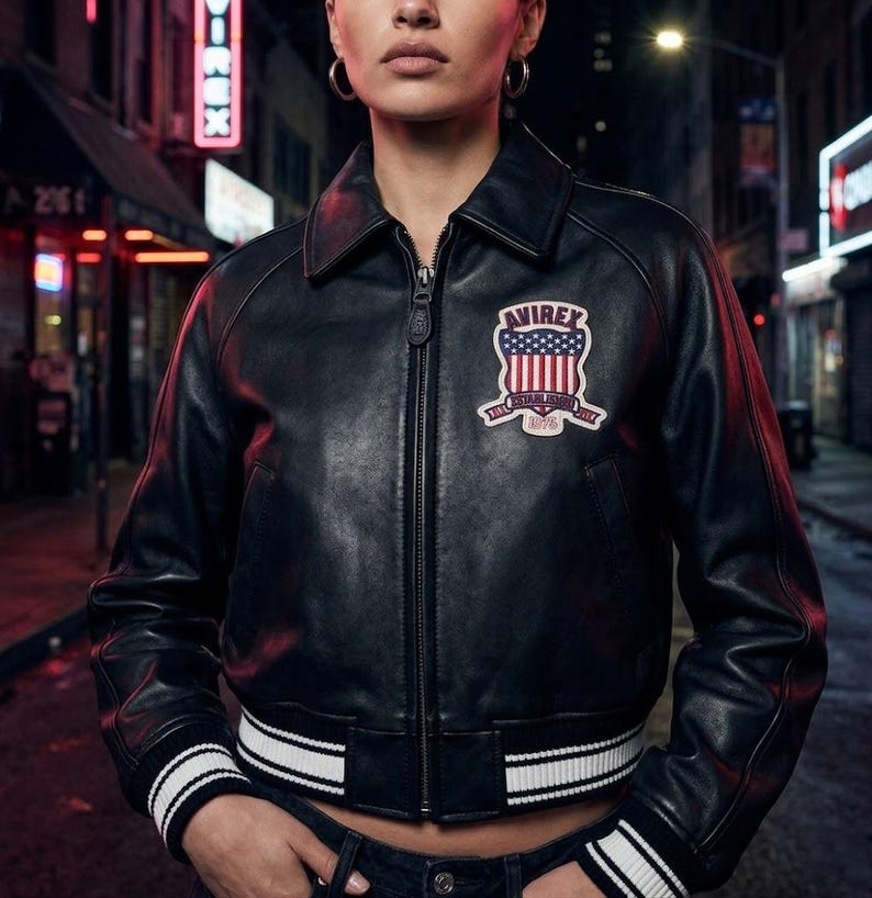 Puede incluir: Chaqueta bomber de cuero negro con cremallera, cuello y ribete de rayas blancas en los pu&ntilde;os y el dobladillo. Cuenta con un parche en el pecho con el texto "AVIREX" y un dise&ntilde;o de bandera estadounidense. La chaqueta es corta.