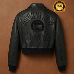Puede incluir: Chaqueta bomber de cuero negro con las palabras "AVIREX" y "USA" en relieve en la espalda. La chaqueta tiene dobladillo y pu&ntilde;os de canal&eacute;, y un cuello. Una pegatina dorada de "BEST SELLER" est&aacute; en la esquina superior derecha.