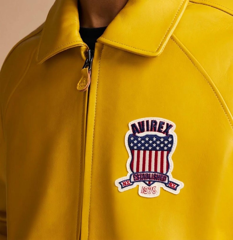 May include: A bright yellow leather jacket with a full-length front zipper and a pointed collar. A patch on the chest features the text AVIREX, ESTABLISHED 1975, and N.Y., along with a shield graphic containing stars and vertical red and white stripes.