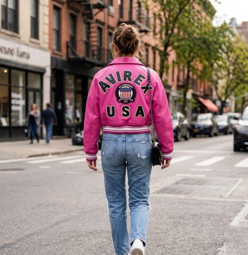 Puede incluir: Una persona lleva una chaqueta de cuero rosa brillante con las palabras "AVIREX" y "USA" en la espalda, adem&aacute;s de un emblema circular. La chaqueta tiene detalles en blanco y negro. La persona lleva vaqueros azules y zapatillas blancas, caminando por una calle.