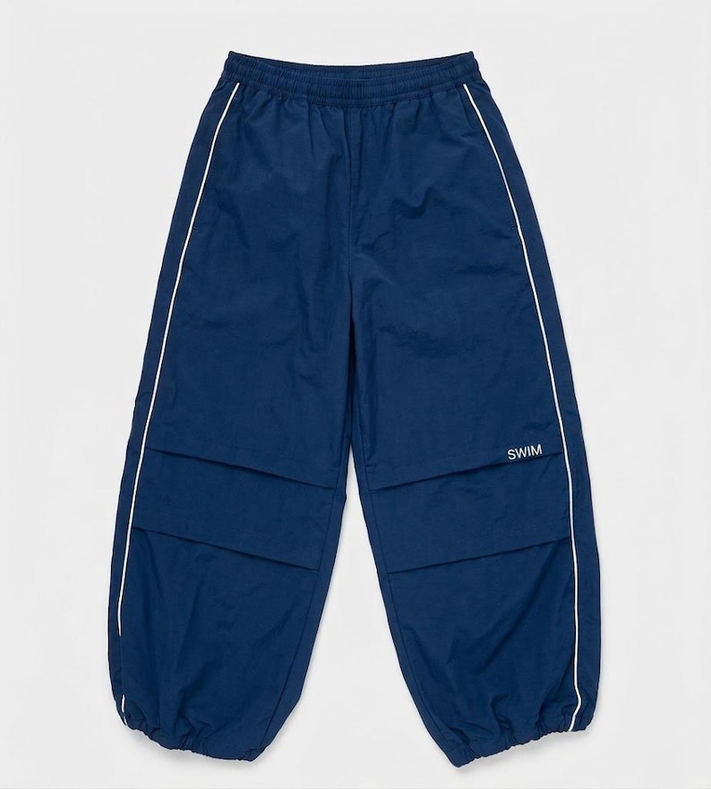 May include: Navy blue athletic pants featuring an elastic waistband, white piping along the side seams, and horizontal paneling across the thighs. The word SWIM is printed in white on the right thigh, and the leg openings have elastic cuffs.