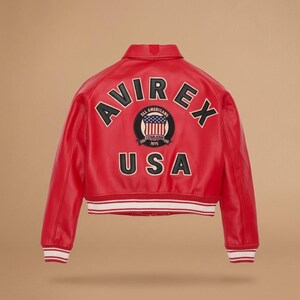 Puede incluir: Chaqueta de cuero roja con la palabra "AVIREX" en letras negras sobre los hombros. Debajo, un parche circular con "ALL AMERICANS" y "USA" en negro. La chaqueta tiene un dobladillo a rayas blancas y rojas. Estilo bomber cl&aacute;sico.