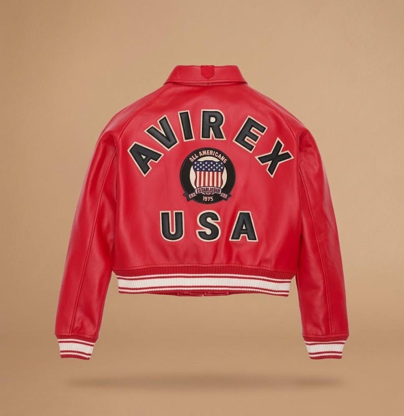 Puede incluir: Chaqueta de cuero roja con la palabra "AVIREX" en letras negras sobre los hombros. Debajo, un parche circular con "ALL AMERICANS" y "USA" en negro. La chaqueta tiene un dobladillo a rayas blancas y rojas. Estilo bomber cl&aacute;sico.