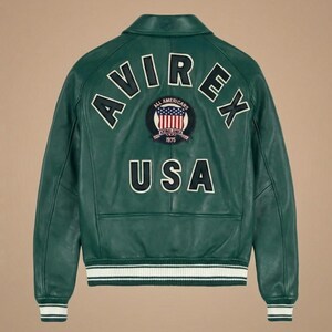 May include: Green leather jacket with white and black trim on the cuffs and waist. The back features the word "AVIREX" in white lettering, along with a patch that says "ALL AMERICANS 1975 USA".