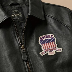 K&ouml;nnte beinhalten: Eine schwarze Lederjacke mit Frontrei&szlig;verschluss und spitzem Kragen. Ein gestickter Aufn&auml;her auf der Brust zeigt den Text AVIREX, ESTABLISHED 1975 und ein Schild-Motiv mit roten und wei&szlig;en Streifen sowie wei&szlig;en Sternen auf blauem Grund.