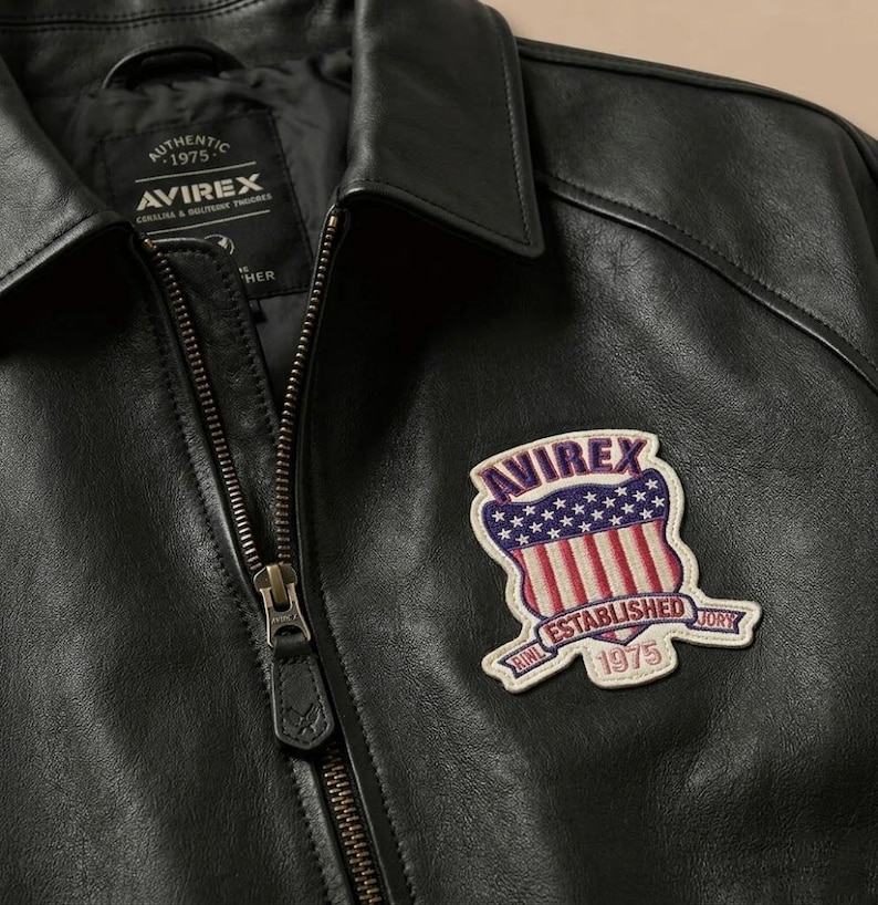 K&ouml;nnte beinhalten: Eine schwarze Lederjacke mit Frontrei&szlig;verschluss und spitzem Kragen. Ein gestickter Aufn&auml;her auf der Brust zeigt den Text AVIREX, ESTABLISHED 1975 und ein Schild-Motiv mit roten und wei&szlig;en Streifen sowie wei&szlig;en Sternen auf blauem Grund.