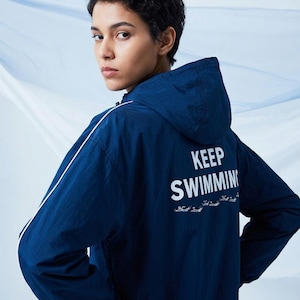 May include: A dark blue hooded windbreaker jacket featuring a white stripe along the sleeves and the text "KEEP SWIMMING" printed on the back in white, accompanied by a small graphic of swimmers below the text.