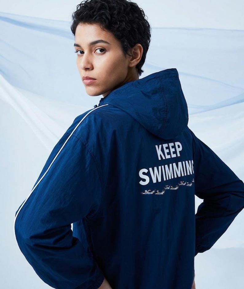 May include: A dark blue hooded windbreaker jacket featuring a white stripe along the sleeves and the text "KEEP SWIMMING" printed on the back in white, accompanied by a small graphic of swimmers below the text.