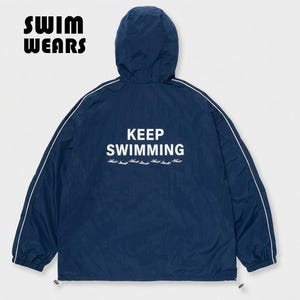 Puede incluir: Una chaqueta cortavientos con capucha de color azul oscuro con ribetes blancos a lo largo de las mangas y la capucha. La espalda muestra el texto blanco "KEEP SWIMMING" sobre una fila de pequeños gráficos de peces blancos. "SWIM WEARS" aparece arriba a la izquierda.