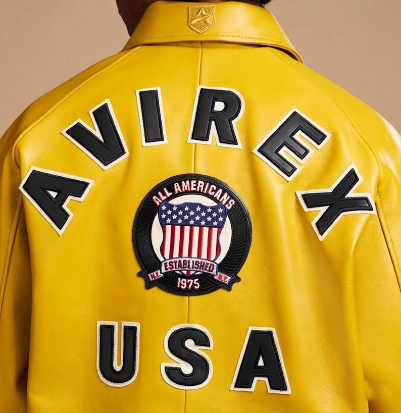 May include: A bright yellow leather jacket featuring the black text AVIREX arched across the upper back. A circular patch in the center displays an American flag shield with the text ALL AMERICANS ESTABLISHED 1975 N.Y. The word USA is in black below.