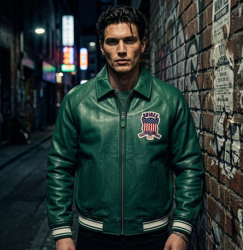 May include: A green leather jacket with a zip-up front and a collar. The jacket has a patch on the chest with the text "AVIREX" and a design of red and blue stripes and stars. The jacket has white stripes on the cuffs and the bottom hem.