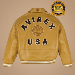 May include: Mustard yellow leather jacket with the text "AVIREX USA" on the back. The jacket has a collar, a logo on the upper back, and white and brown striped cuffs and hem. A gold seal reads "ORIGINAL PRODUCT BEST SELLER BEST QUALITY".