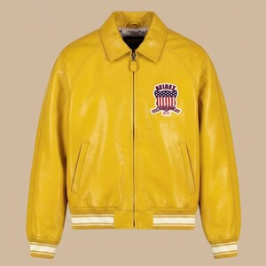 May include: A bright yellow leather jacket with a full-length front zipper, a classic collar, and ribbed cuffs and hem featuring white stripes. On the left chest, an embroidered patch displays the text AVIREX with a red, white, and blue flag design.