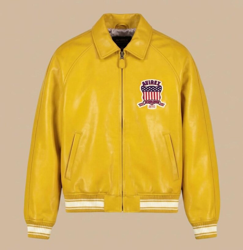 May include: A bright yellow leather jacket with a full-length front zipper, a classic collar, and ribbed cuffs and hem featuring white stripes. On the left chest, an embroidered patch displays the text AVIREX with a red, white, and blue flag design.