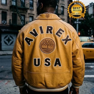 May include: Mustard yellow leather jacket with the word "AVIREX" arched across the back, above "USA". A circular patch with a tree design is centered below the brand name. The jacket has a white and black striped trim at the bottom.