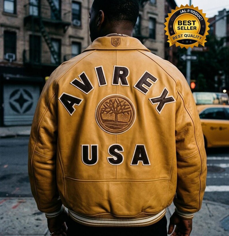 May include: Mustard yellow leather jacket with the word "AVIREX" arched across the back, above "USA". A circular patch with a tree design is centered below the brand name. The jacket has a white and black striped trim at the bottom.