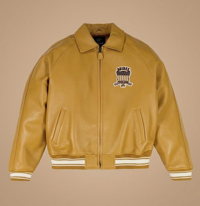 May include: Mustard yellow leather jacket with a zip closure, collar, and ribbed cuffs and hem. Features a patch on the chest with the text "AVIREX 1975". The jacket has two front pockets and a classic bomber jacket design.