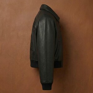 Puede incluir: Chaqueta bomber de cuero negro con cuello y pu&ntilde;os acanalados. La chaqueta tiene cierre de cremallera y un peque&ntilde;o bolsillo en la parte delantera. La chaqueta se muestra sobre un fondo marr&oacute;n.