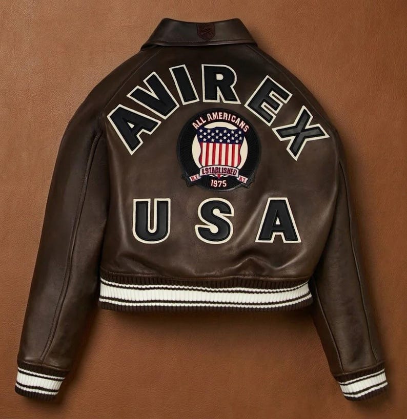 Puede incluir: Chaqueta de cuero marr&oacute;n con letras y parches blancos y negros. La parte trasera de la chaqueta presenta la palabra "AVIREX" arqueada en la parte superior, con "USA" debajo. Un parche circular con la bandera americana en el centro.