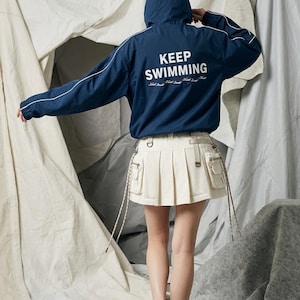 May include: A dark blue hooded windbreaker jacket with white piping on the sleeves and the text KEEP SWIMMING printed in white on the back, paired with a cream-colored pleated mini skirt featuring side cargo pockets and hanging drawstrings.