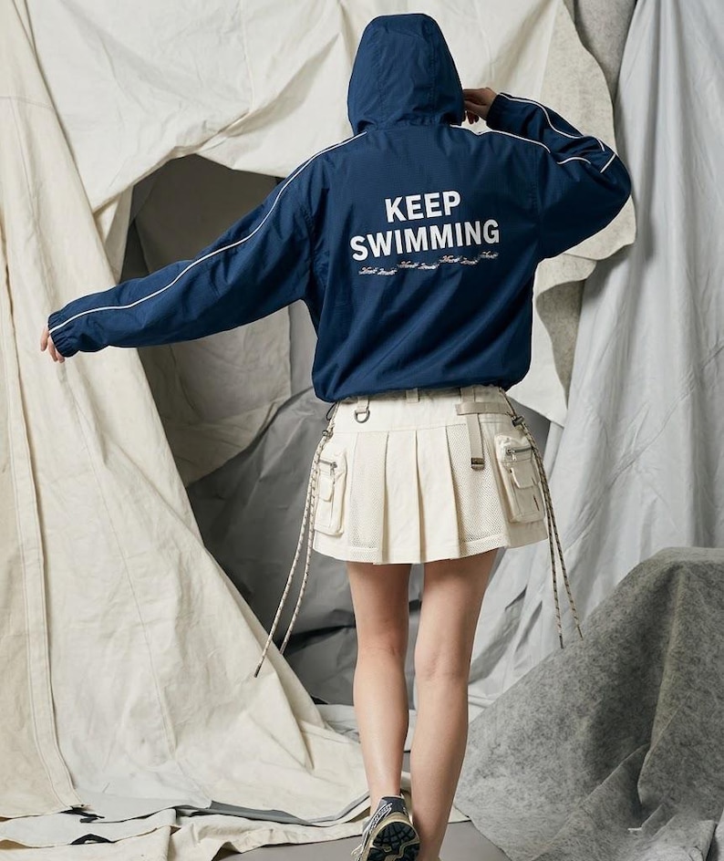 May include: A dark blue hooded windbreaker jacket with white piping on the sleeves and the text KEEP SWIMMING printed in white on the back, paired with a cream-colored pleated mini skirt featuring side cargo pockets and hanging drawstrings.