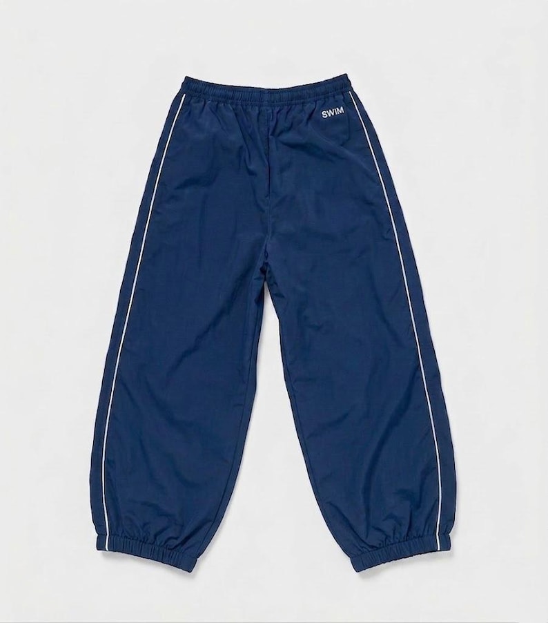 May include: Navy blue athletic pants featuring an elastic waistband and cuffs. The pants have a white vertical piping detail running down the outer legs and the word SWIM printed in white text on the upper right side near the waistband.