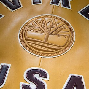 May include: Close-up of a mustard-colored leather jacket with a large, circular embroidered patch featuring a tree design. The patch is surrounded by a rope-like border. Brown and white lettering is visible.