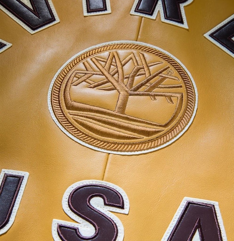 May include: Close-up of a mustard-colored leather jacket with a large, circular embroidered patch featuring a tree design. The patch is surrounded by a rope-like border. Brown and white lettering is visible.