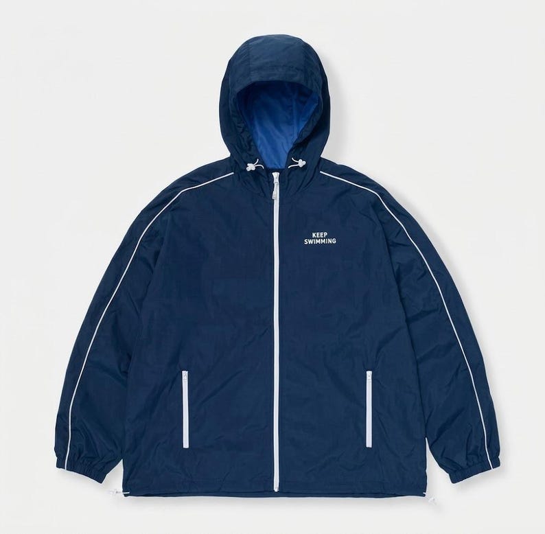 May include: A navy blue windbreaker jacket with a hood, featuring a white front zipper, white piping along the sleeves, and two side pockets. The text KEEP SWIMMING is printed in white on the upper left chest, and the interior lining is bright blue.