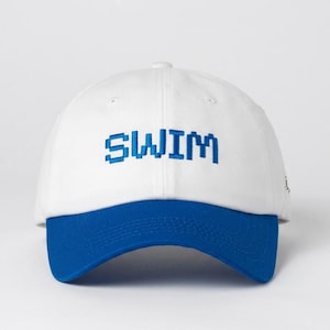 May include: A white baseball cap with a structured crown and a solid blue brim. The word SWIM is embroidered across the front center in a blue, pixelated, block-style font. The cap features a button on top and stitched eyelets for ventilation.